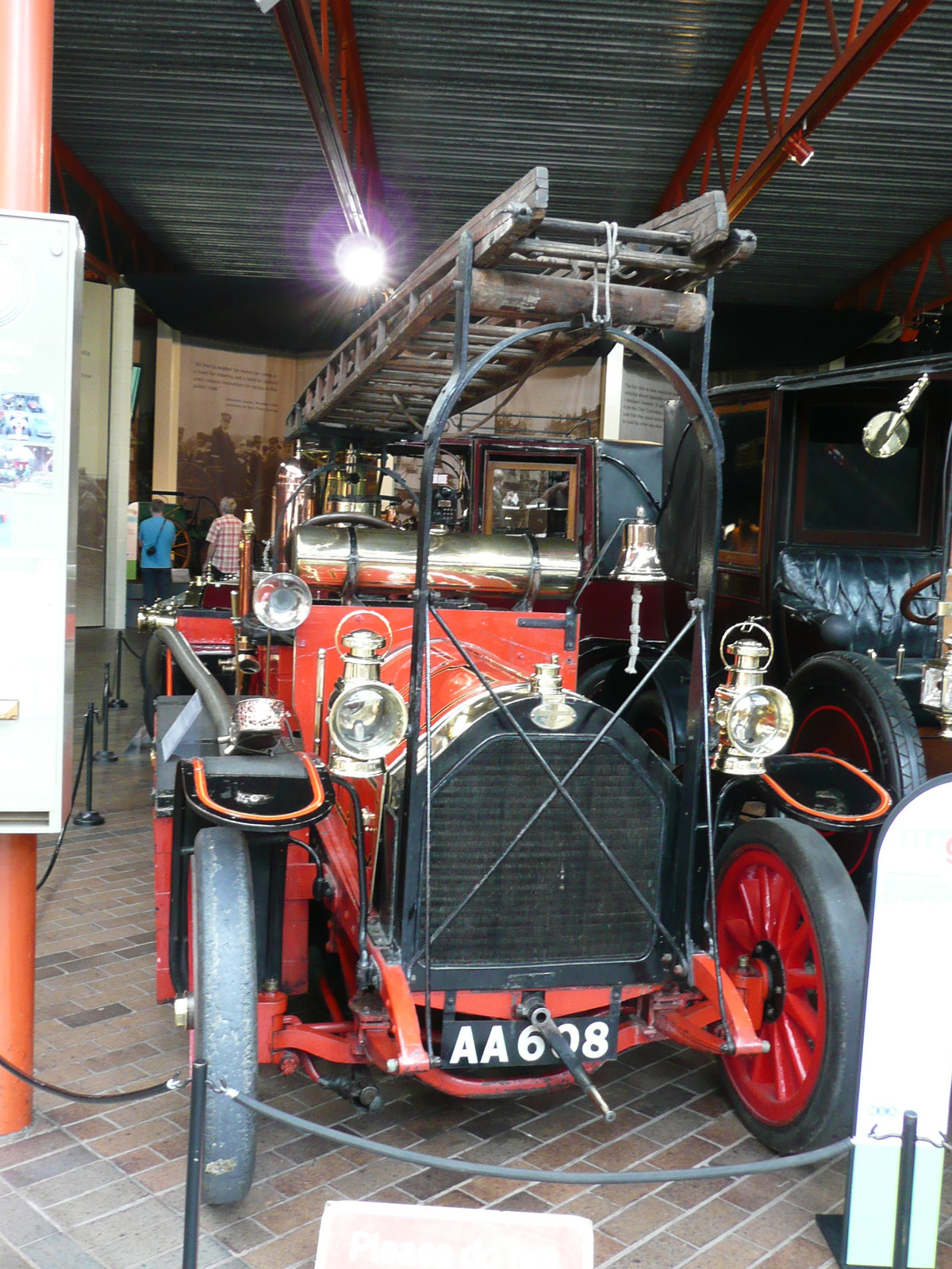 Frontal view of a red and black period fire appliance, brass lamps, radiator, solid front tyres and overhead ladder. Museum surroundings, ropes, signs and glare remain. Matches the named Beaulieu converted vehicle; does not establish serial numbers or engine internals. Not calibrated geometry evidence. Whole image resized and JPEG re-encoded; original is public domain.