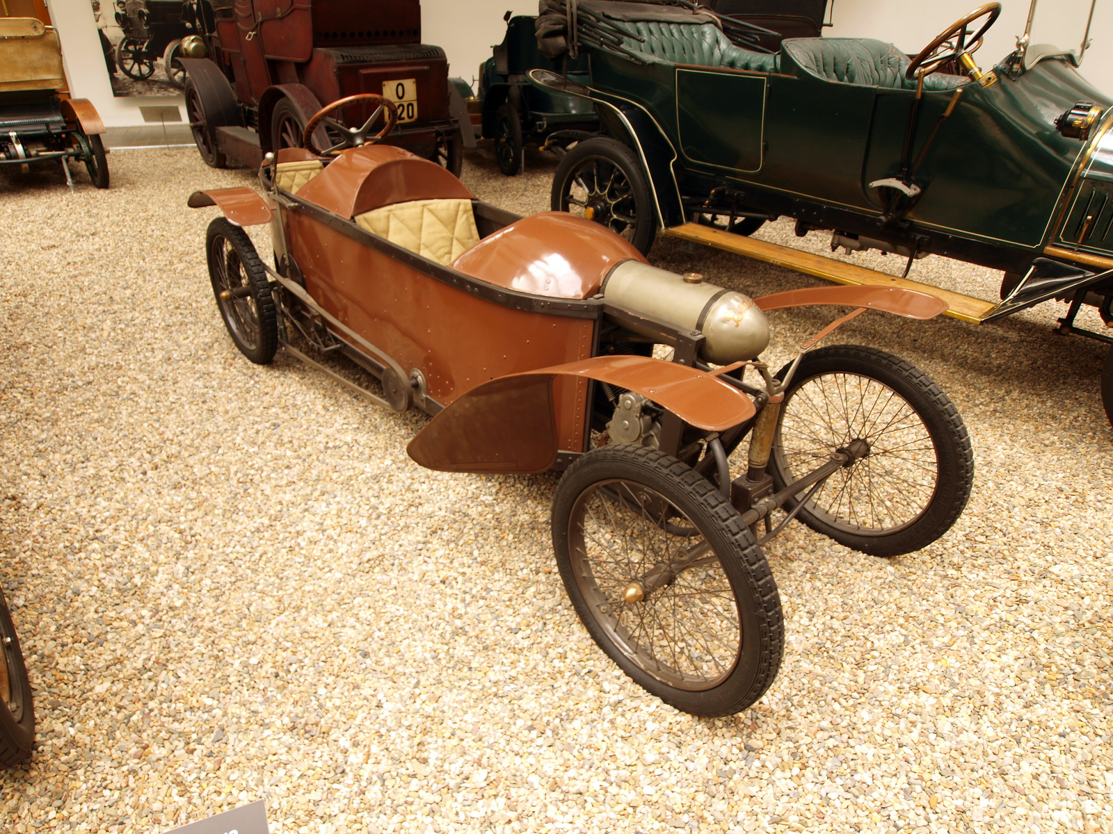 Brown narrow tandem-seat cyclecar with rear steering wheel, exposed belt drive, spoked wheels and separate curved mudguards, viewed from the front three-quarter angle in a museum. The whole vehicle is legible; neighboring exhibits and reflections remain. Original and derivative show the same unaltered composition. No readable chassis number or engine identity; acceptable only as a disclosed representative BD2 cover, not exact-chassis or calibrated geometry evidence.