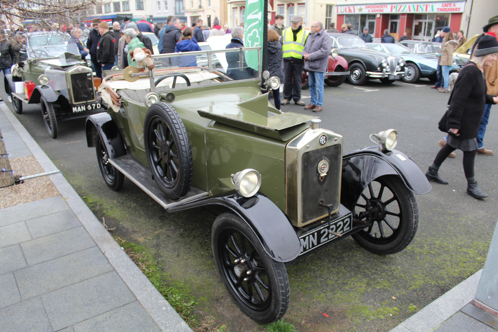 Green ordinary A.B.C. touring car with upright full-width windscreen, side door, spare wheel, opened bonnet and readable ABC badge/MN 2222 plate. Car body and all visible wheels remain legible. Original and 1600×1067 derivative have the same whole-image composition, with spectators and other cars in the background. No chassis/year proof or exact match to Lane's red survivor; accepted only as a clearly disclosed representative.