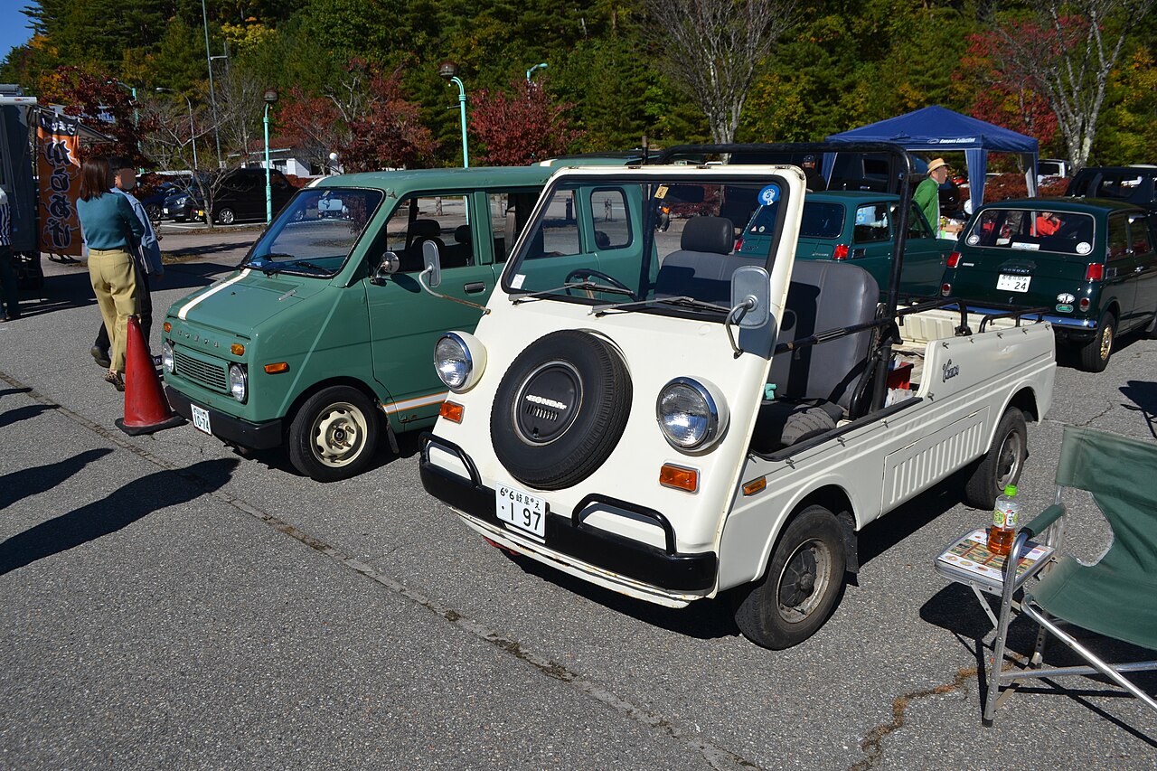 White foreground vehicle: two-seat Vamos Honda 2, photographed in 2023. Exact build year and originality unverified; representative of the 1970 launch type. The green van beside it is a different Honda Life Step Van.