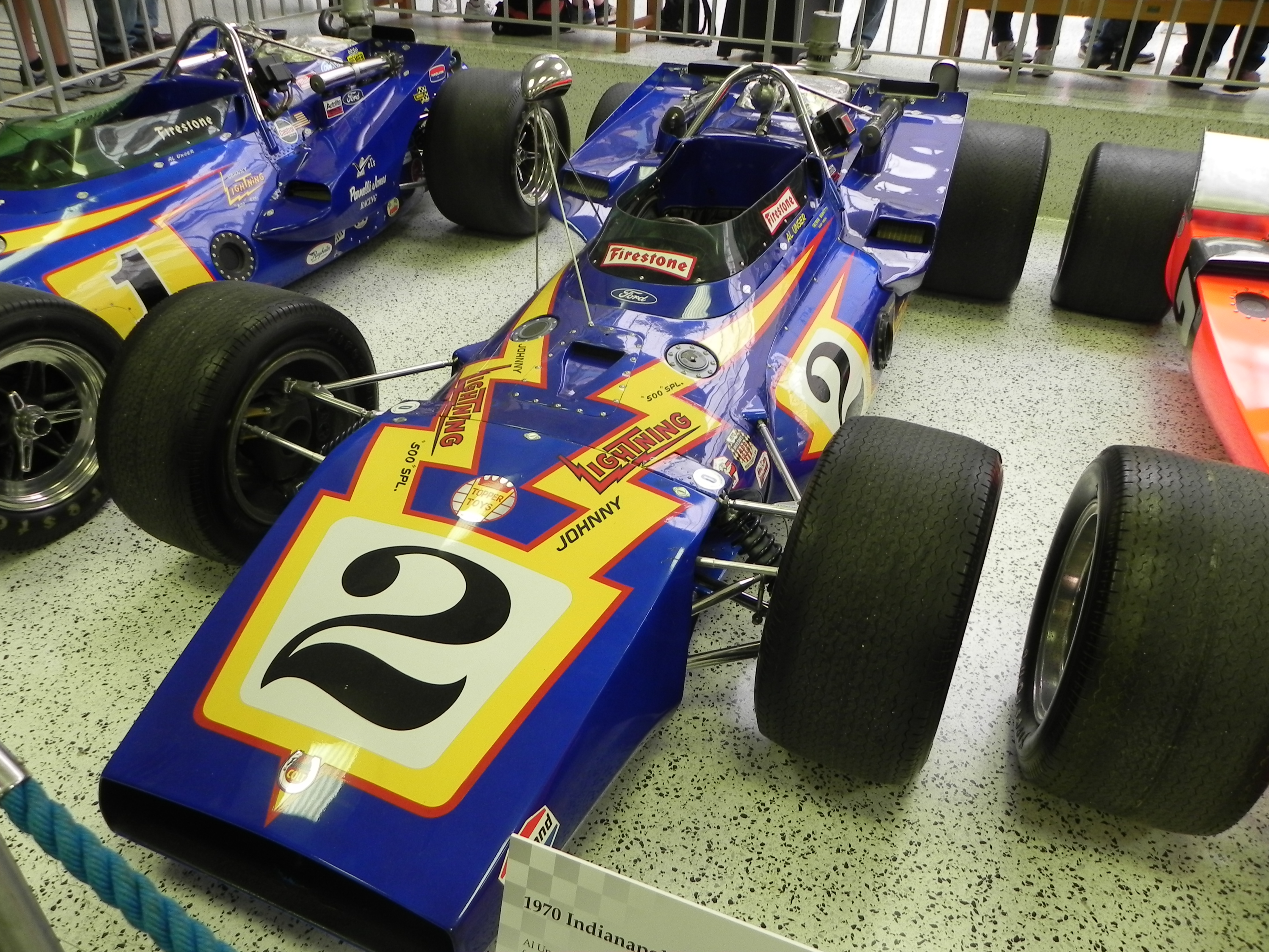 The actual blue no. 2 Johnny Lightning P.J. Colt/Ford displayed at the Indianapolis Motor Speedway Museum, viewed from the front left with neighboring historic race cars visible.