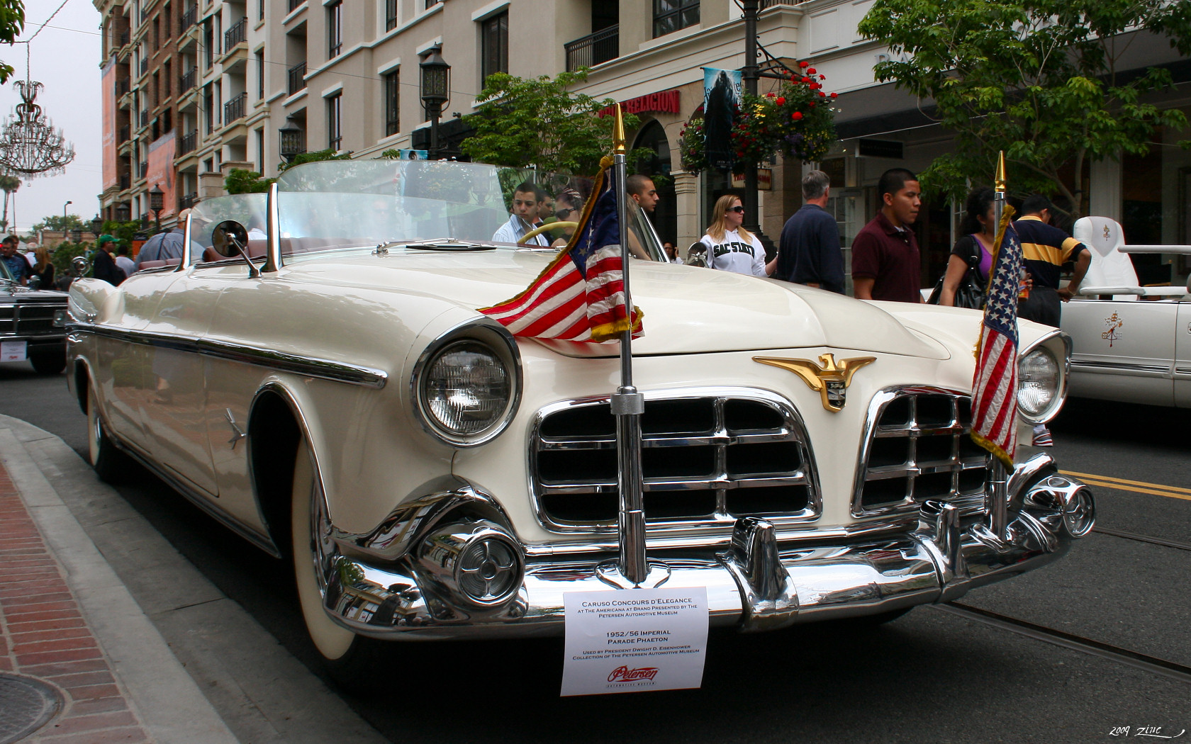 White open Chrysler Imperial Parade Phaeton viewed from the front right, with two flags and a Petersen museum placard identifying Eisenhower use.