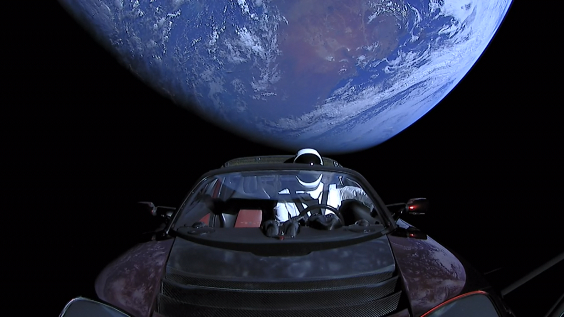 Elon Musk’s actual first-generation Tesla Roadster floating in space with the Starman mannequin at the wheel and Earth behind it; no registration is visible.
