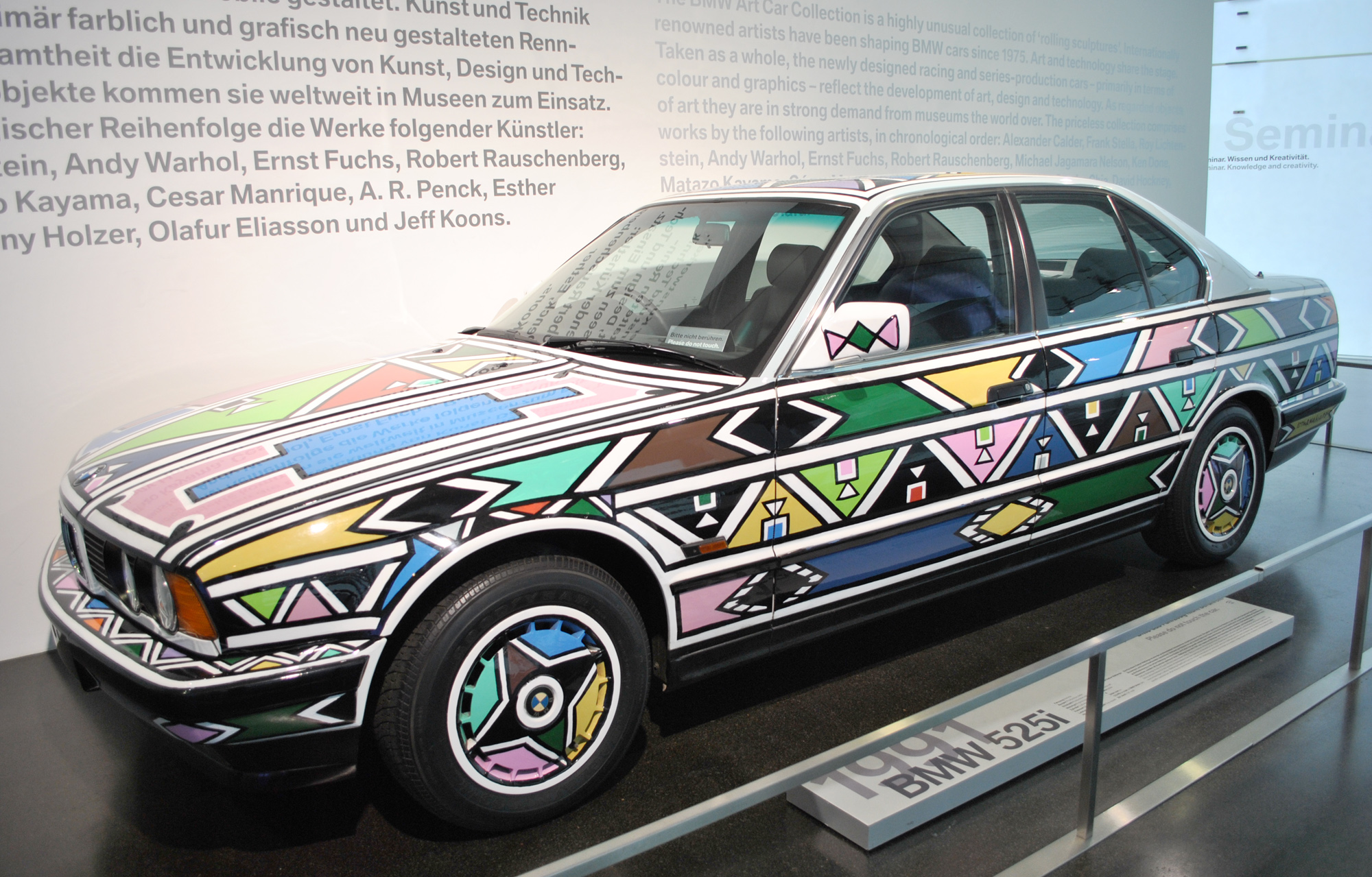 Esther Mahlangu’s actual 1991 BMW 525i Art Car displayed at the BMW Museum, covered in bright geometric Ndebele patterns; no registration is visible.