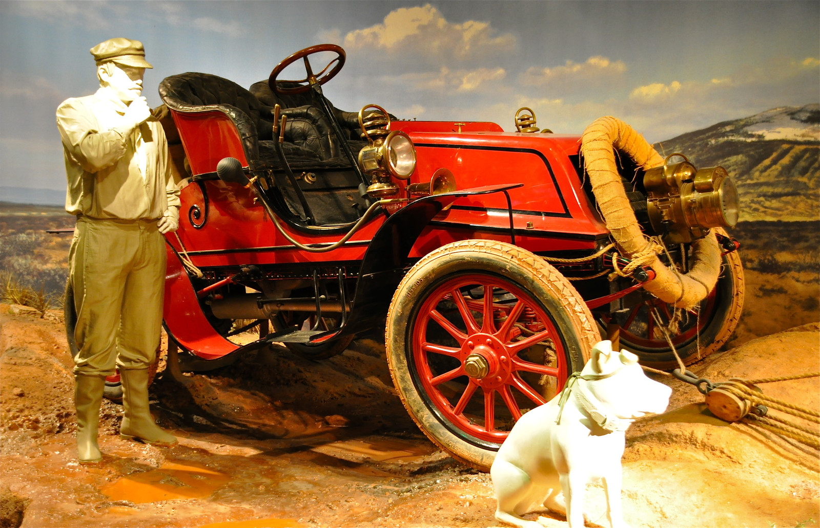 Red 1903 Winton touring car “Vermont” displayed with museum figures of Horatio Nelson Jackson and his dog Bud in a constructed landscape scene.
