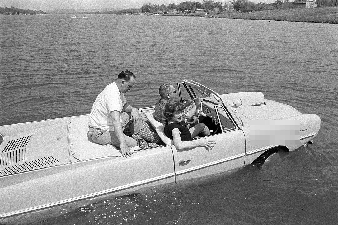 President Lyndon B. Johnson driving an Amphicar in a ranch lake with three passengers in 1965; its historic registration is pixelated.