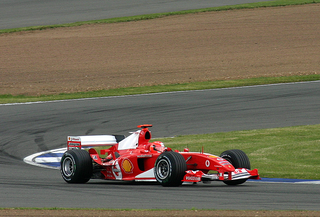 Michael Schumacher driving the red race-number-1 Ferrari F2004 through Brooklands at Silverstone on July 10, 2004; no registration is visible.