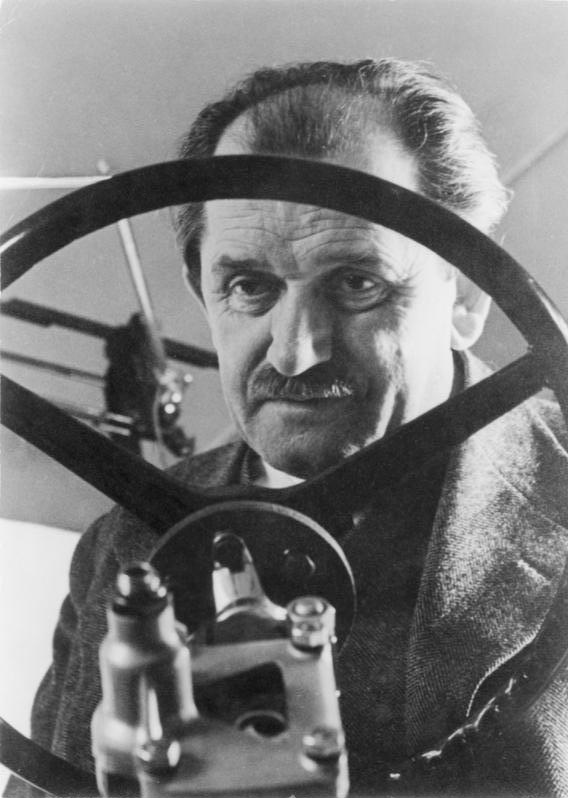 Black-and-white 1940 portrait of Ferdinand Porsche framed by a steering wheel.