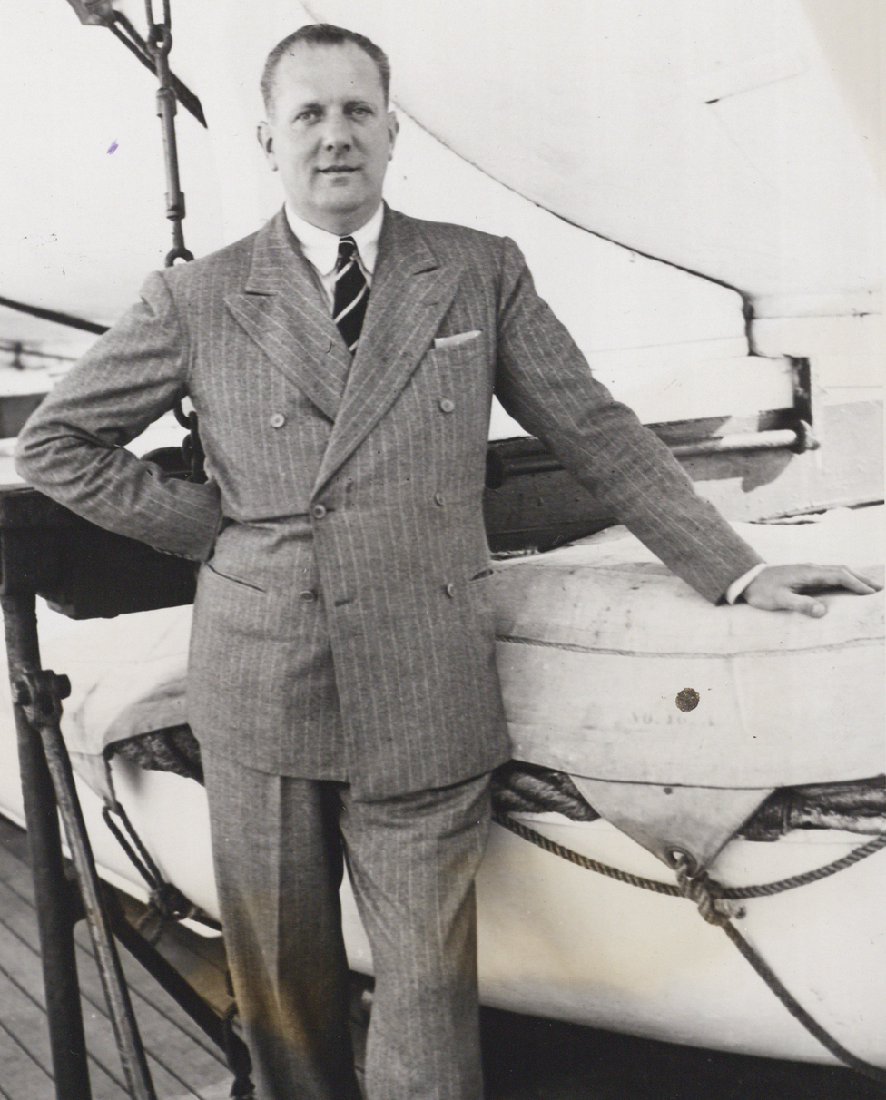Black-and-white 1935 portrait of racing driver John Cobb in a double-breasted suit standing beside a boat.