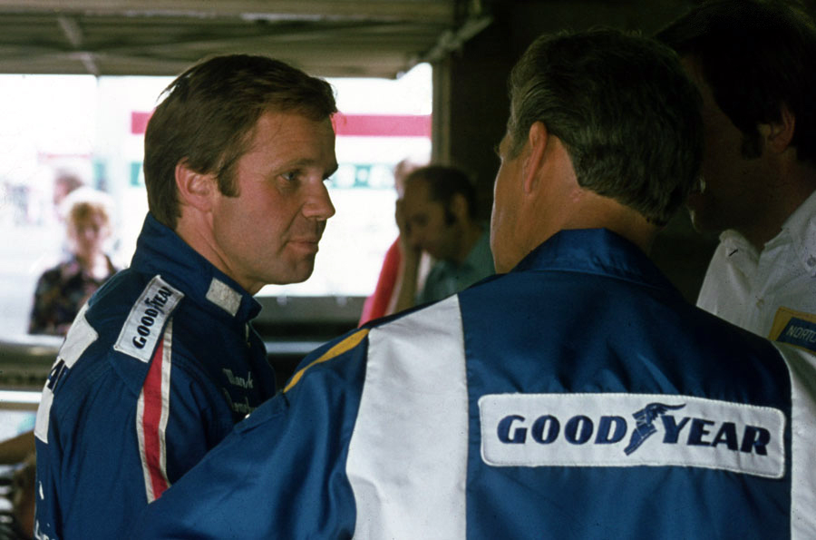 Mark Donohue in a blue racing suit speaking with team members at the 1975 British Grand Prix, photographed by Gillfoto.