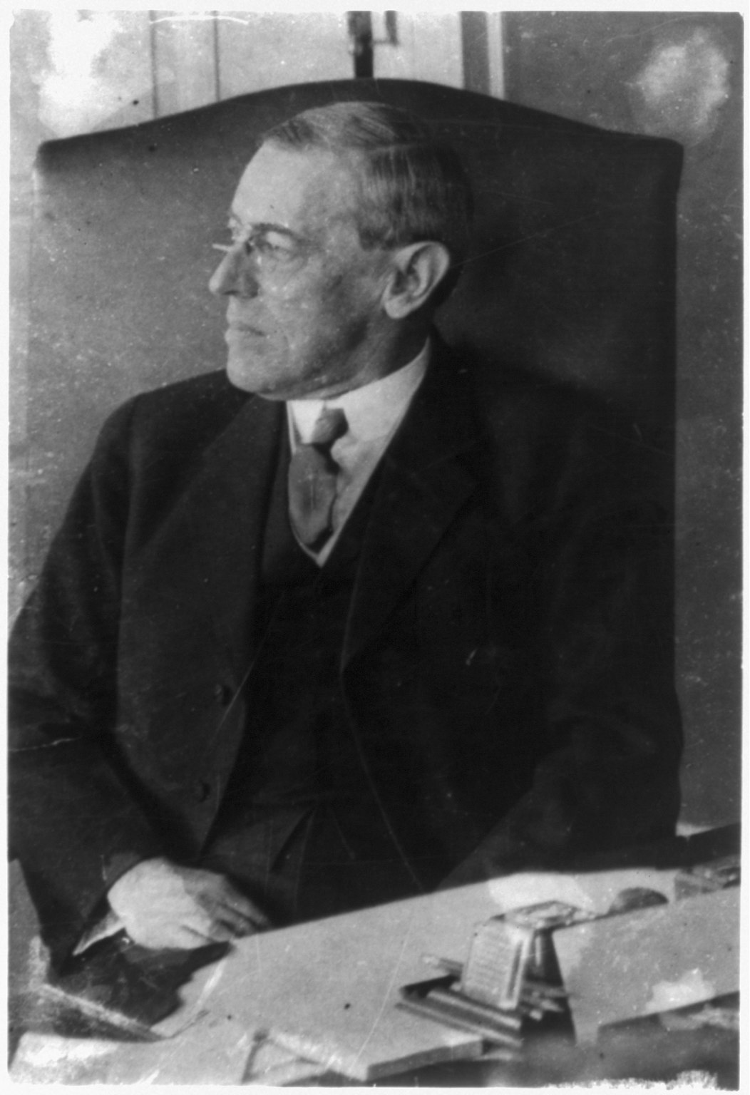 Black-and-white 1913 portrait of Woodrow Wilson seated at a desk, facing left.