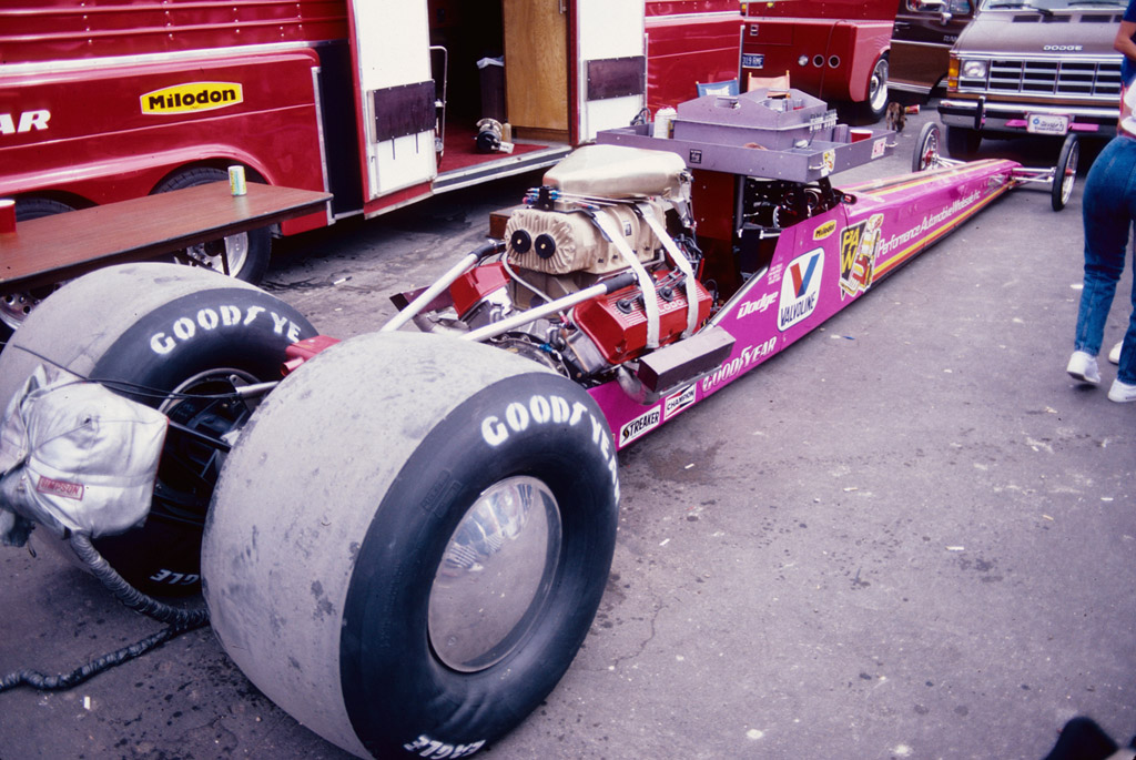 Shirley Muldowney’s actual pink Top Fuel dragster at the 1986 NHRA Chief Auto Parts Nationals, viewed from the front and side in the event paddock; no registration is visible.