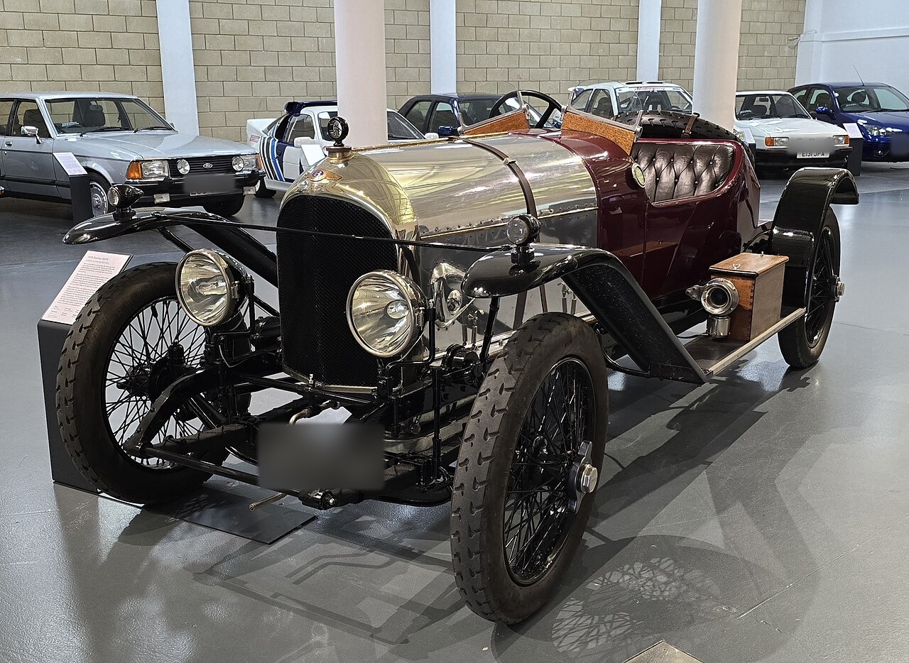 The actual rebuilt Bentley EXP2 displayed indoors, viewed from the front left with its polished bonnet, dark-red body and four spoked wheels visible; all readable registration plates are blurred.